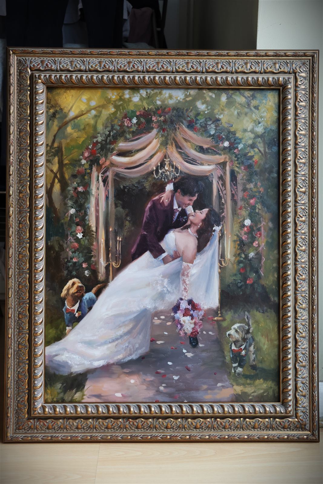 Finished wedding oil painting in an ornate gold frame: the couple beneath a floral arch, the groom in burgundy, their two dogs at their feet