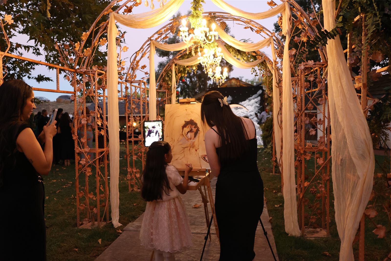 Caitlin painting live at her easel beneath a floral arch as guests watch at dusk